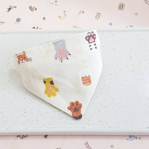 K&ouml;nnte beinhalten: Wei&szlig;es Baumwollbandana mit Pfotenabdruck- und Katzendesign. Das Bandana hat verschiedene Pfotenabdr&uuml;cke in Orange, Braun und Grau.