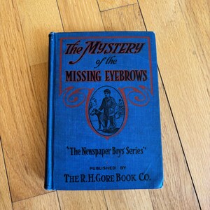 May include: A vintage blue book titled "The Mystery of the Missing Eyebrows" with red lettering. The cover features an illustration of a person with a dog. Published by The R.H. Gore Book Co.