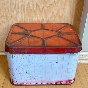 May include: A rectangular metal storage tin with a red and orange lid and a white and red grid pattern on the sides. The tin has a distressed, vintage appearance, suggesting age and use. The lid features a diamond pattern.