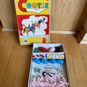 May include: A vintage Cootie game box with a yellow, red, and white design. The box reads "THE GAME OF... COOTIE" and includes game pieces like pink legs, blue body parts, and red heads. The game is described as an "EXCITING EDUCATIONAL GAME FOR ALL AGES."
