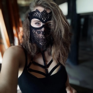 May include: A black lace mask with a floral design covers the face. The mask has eye cutouts and extends down to cover the nose and mouth. A black tank top and harness are also visible.