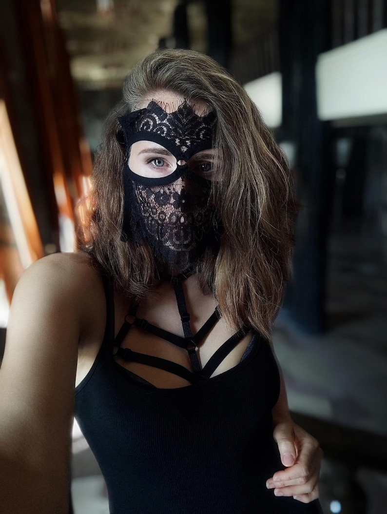 May include: A black lace mask with a floral design covers the face. The mask has eye cutouts and extends down to cover the nose and mouth. A black tank top and harness are also visible.