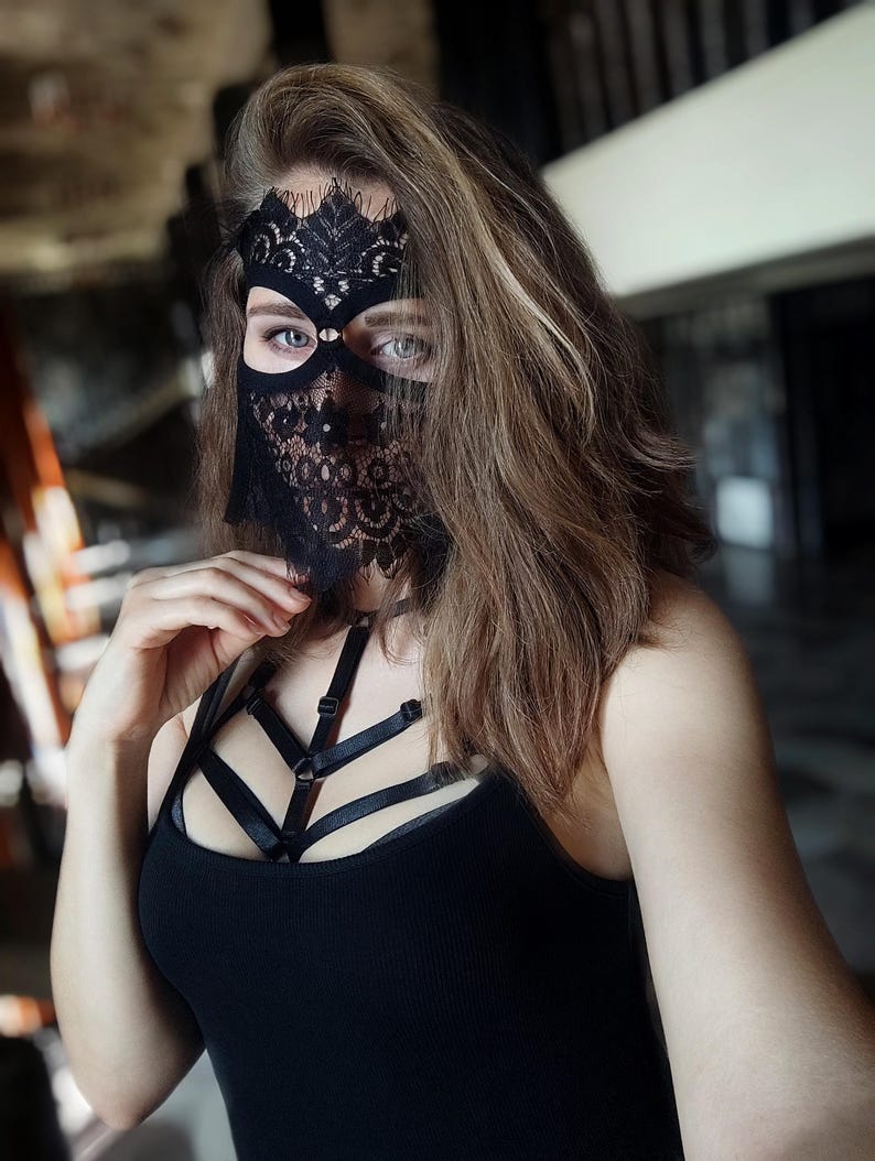 May include: A black lace mask with a floral design covers the eyes and nose. The mask is worn with a black tank top and a black harness. The person has long, light brown hair.