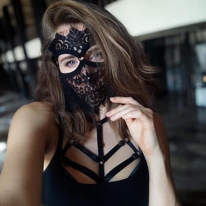 May include: Black lace mask with intricate floral patterns, covering the eyes and lower face. The mask is paired with a black tank top and a harness-style chest piece. The overall aesthetic is dark and sensual.