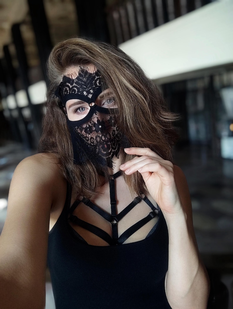 May include: Black lace mask with intricate floral patterns, covering the eyes and lower face. The mask is paired with a black tank top and a harness-style chest piece. The overall aesthetic is dark and sensual.
