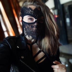 May include: Black lace mask with a delicate floral pattern, covering the eyes and lower face. The mask is worn with a black leather jacket and a black top. The overall aesthetic is dark and mysterious.