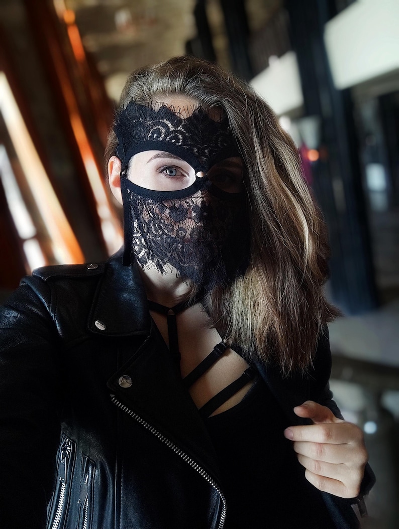 May include: Black lace mask with a delicate floral pattern, covering the eyes and lower face. The mask is worn with a black leather jacket and a black top. The overall aesthetic is dark and mysterious.