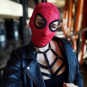 Puede incluir: Una persona lleva una capucha roja estampada con agujeros negros para los ojos y una chaqueta de cuero negra. La persona también lleva un arnés negro con correas y anillos.