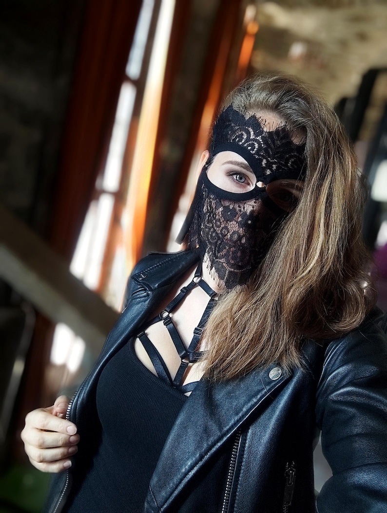 May include: A black lace mask covers the face, paired with a black top and a black leather jacket. The mask has intricate floral patterns. The jacket is zipped up, and the overall look is dark and edgy.