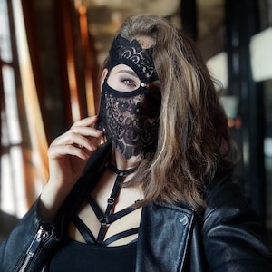 May include: A black lace mask with a floral design covers the eyes and lower face. The person is wearing a black leather jacket and a black top with a harness. The background is blurred, with hints of a window and interior.