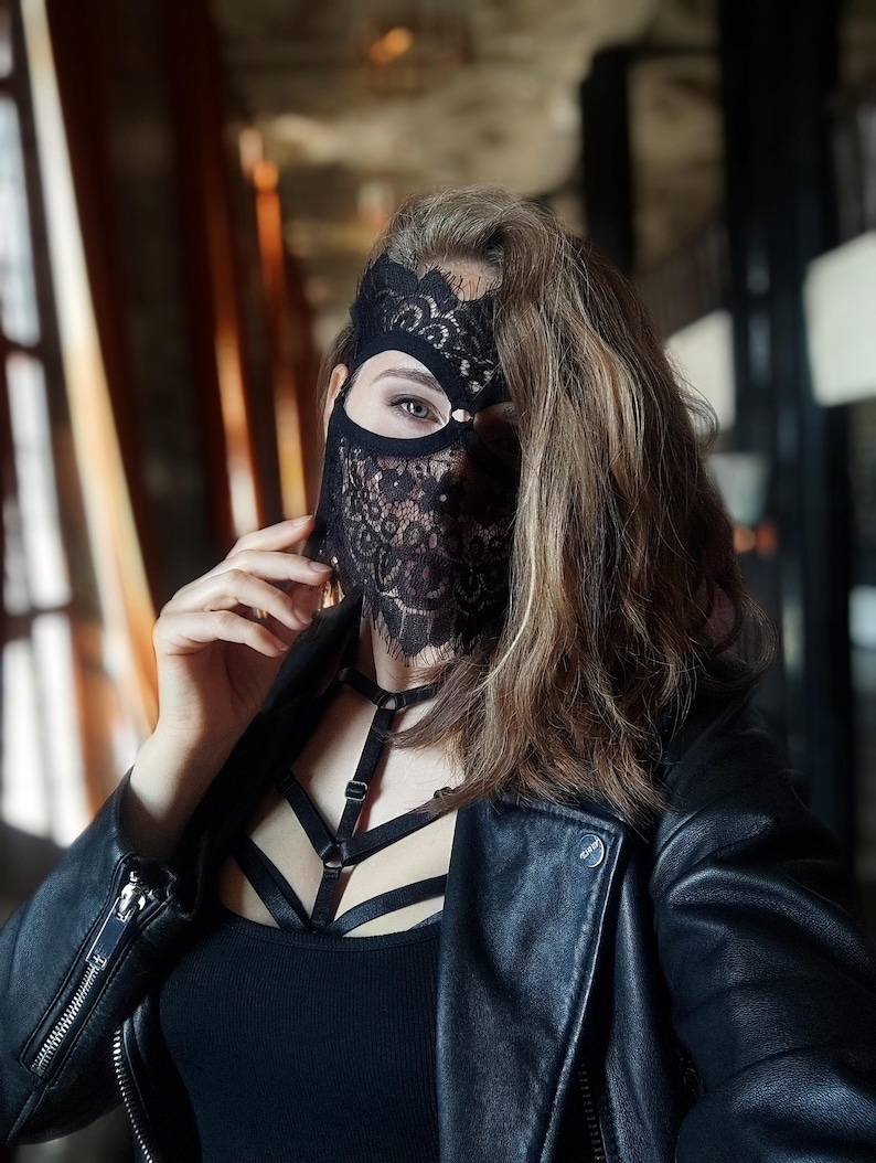 May include: A black lace mask with a floral design covers the eyes and lower face. The person is wearing a black leather jacket and a black top with a harness. The background is blurred, with hints of a window and interior.