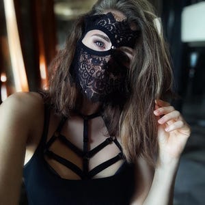 May include: Black lace mask with a floral design covering the eyes and lower face. The mask is paired with a black tank top and a harness-style top. The image is a close-up shot.