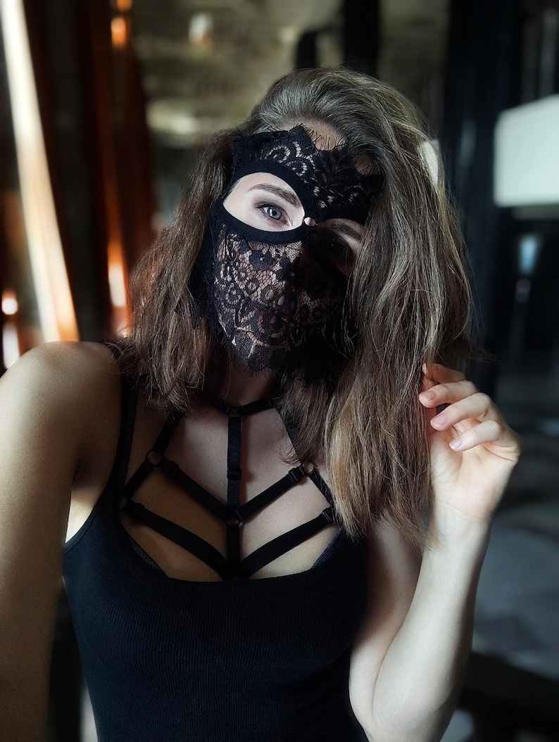 May include: Black lace mask with a floral design covering the eyes and lower face. The mask is paired with a black tank top and a harness-style top. The image is a close-up shot.