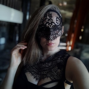 Può includere: Una donna con una maschera di pizzo nero sugli occhi e un top di pizzo nero. La maschera copre tutto il viso tranne la bocca e il mento.