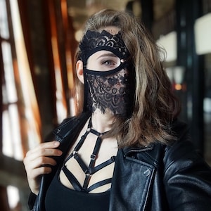 May include: A black lace mask with a floral design covers part of the face. The person is wearing a black leather jacket and a black top with a harness-style design. The background is blurred.