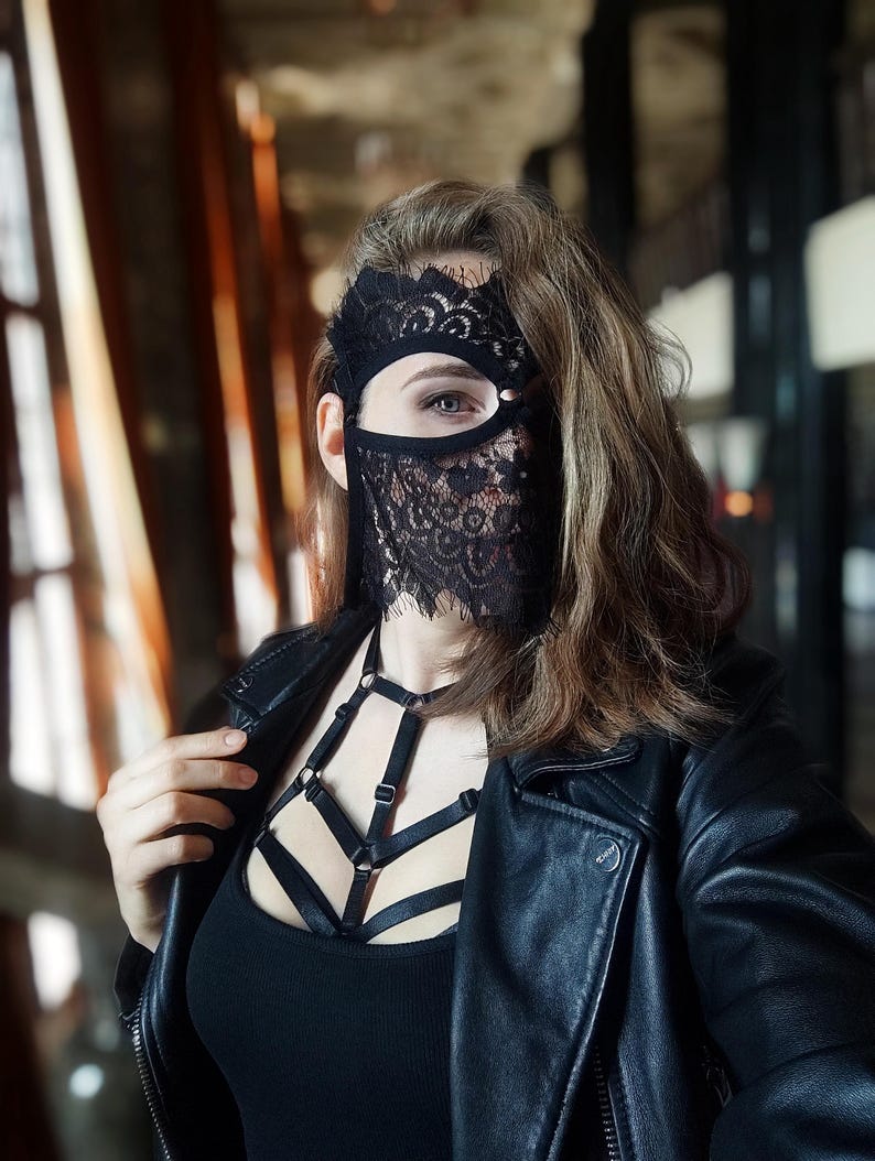 May include: A black lace mask with a floral design covers part of the face. The person is wearing a black leather jacket and a black top with a harness-style design. The background is blurred.