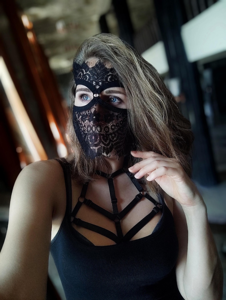 May include: Black lace mask with intricate floral patterns, covering the eyes and lower face. The mask is paired with a black tank top featuring a strappy harness design. The overall aesthetic is dark and mysterious.