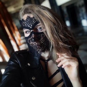 May include: A black lace mask with an intricate floral design covers the eyes and lower face. The mask is worn with a black leather jacket and a black top. The image is a close-up shot, with the focus on the mask and the person's face.