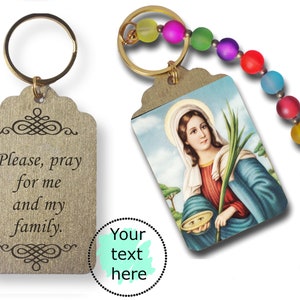 May include: Two gold keychains with a religious theme. One keychain has a wooden tag with the text "Please, pray for me and my family." The other keychain has a small image of a woman holding a branch and a bowl. The keychain also has a rainbow-coloured beaded chain.