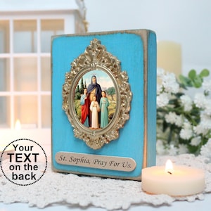 May include: A blue wooden plaque with a gold ornate frame surrounding a picture of Saint Sophia and her three daughters. The plaque has the text "St. Sophia, Pray For Us." on the bottom.