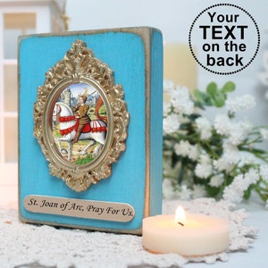 Könnte beinhalten: Ein blauer Holzblock mit einem goldenen verzierten Rahmen, der ein Vintage-Bild von Jeanne d'Arc zu Pferd umrahmt. Der Block hat eine kleine Plakette unten, auf der "St. Jeanne d'Arc, bete für uns" steht.