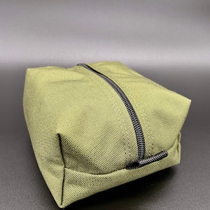 May include: Olive green rectangular pouch with a black zipper closure. The pouch is made of a textured fabric and has a simple, minimalist design. The zipper runs across the top of the pouch.