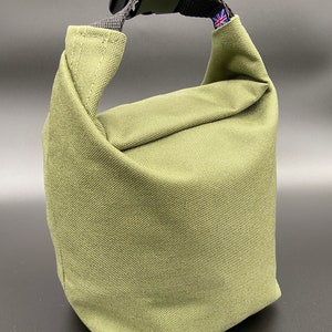 May include: Olive green lunch bag with a black strap and buckle closure. The bag is made of a durable, textured fabric. A small Union Jack flag is visible on the strap. The bag is designed to be folded over and secured with the buckle.