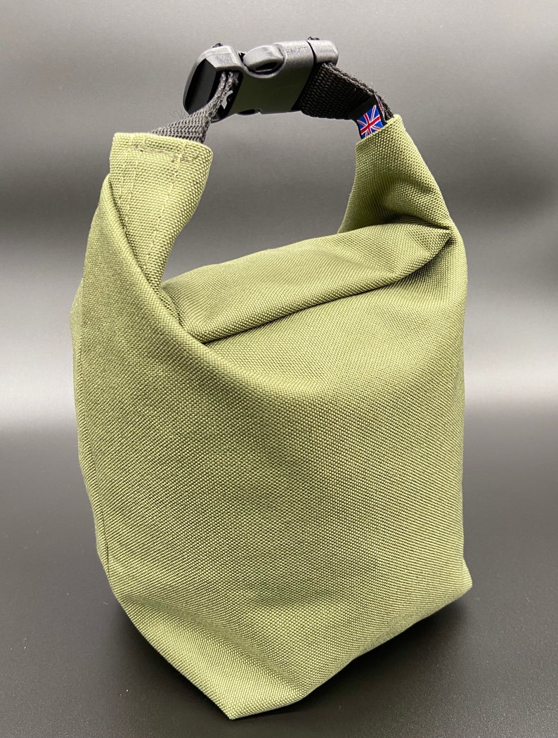 May include: Olive green lunch bag with a black strap and buckle closure. The bag is made of a durable, textured fabric. A small Union Jack flag is visible on the strap. The bag is designed to be folded over and secured with the buckle.