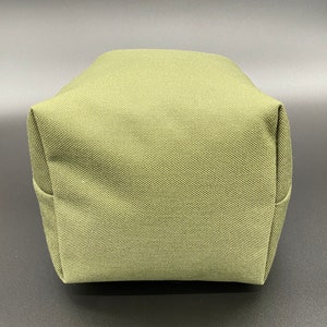 May include: A close-up of a small, olive-green, rectangular bag. The bag is made of a textured fabric and has a boxy shape with folded corners. The bag is sitting on a dark surface.