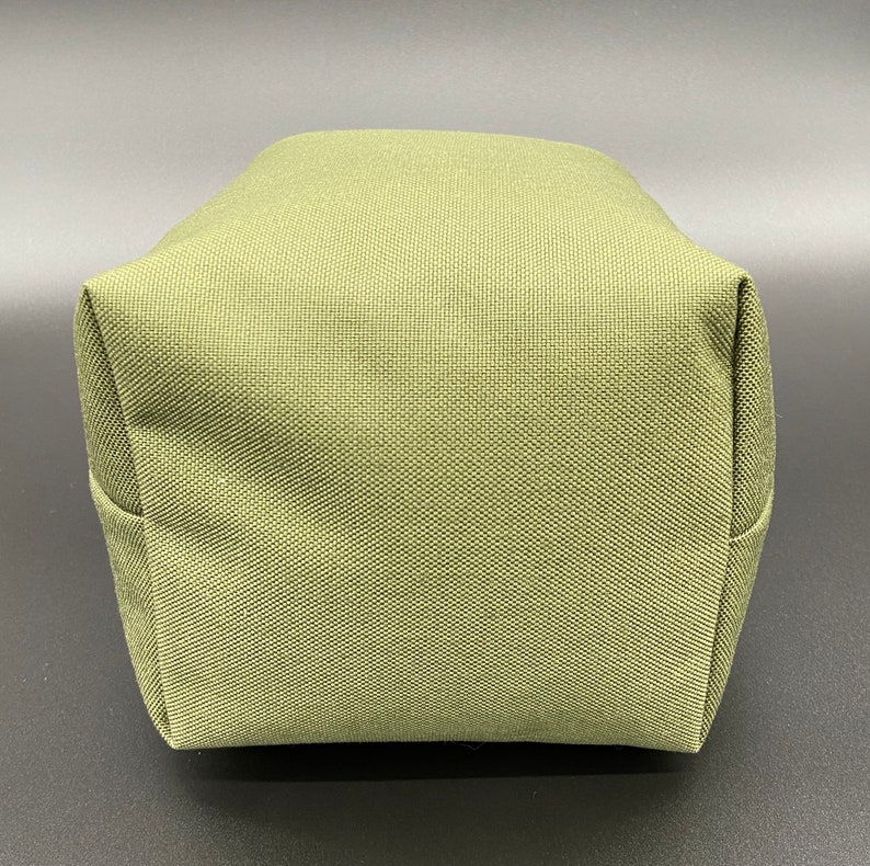 May include: A close-up of a small, olive-green, rectangular bag. The bag is made of a textured fabric and has a boxy shape with folded corners. The bag is sitting on a dark surface.