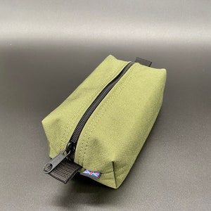 Puede incluir: Bolsa rectangular verde oliva con cremallera negra y tirador. La bolsa tiene un pequeño lazo de tela negra. Es visible una pequeña etiqueta con la bandera británica.