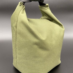 May include: Olive green lunch bag with a black buckle closure and handle. The bag is made of a textured fabric and has a rectangular shape with a folded top. The handle is made of black nylon.