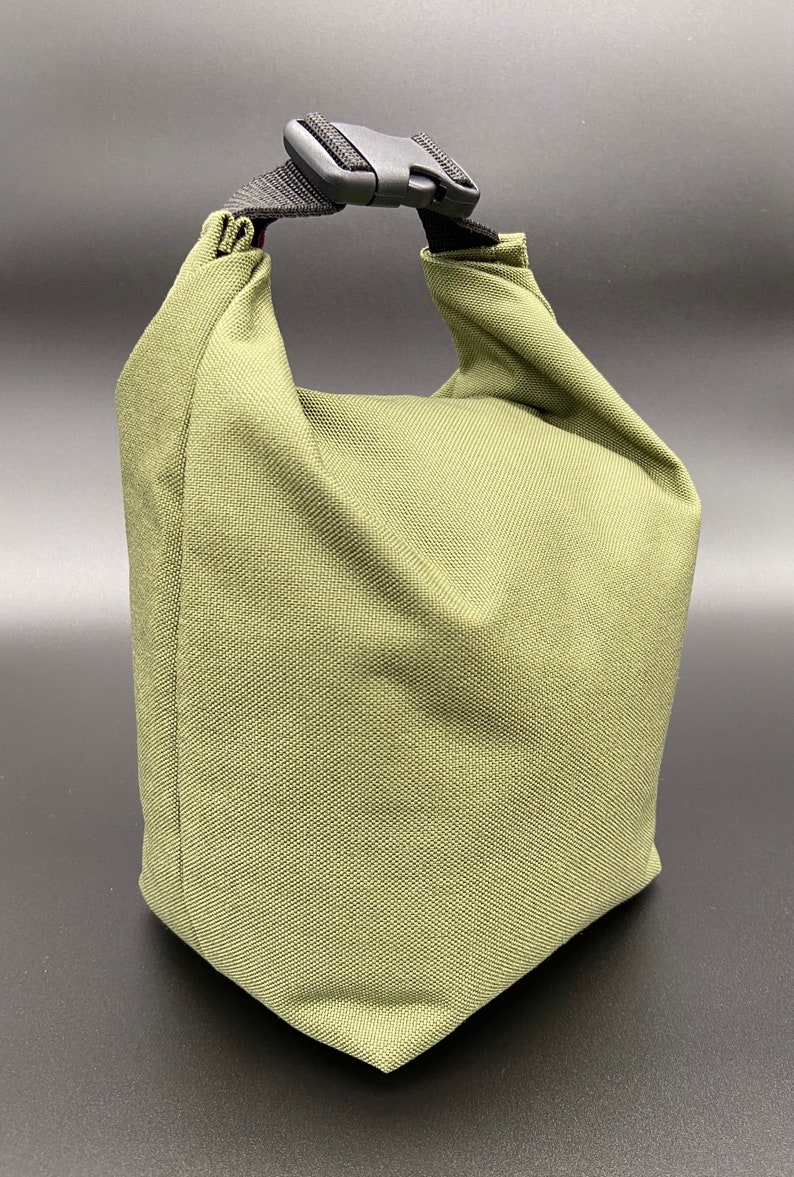 May include: Olive green lunch bag with a black buckle closure and handle. The bag is made of a textured fabric and has a rectangular shape with a folded top. The handle is made of black nylon.