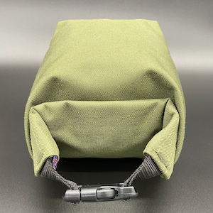 May include: Olive green rectangular pouch with a black buckle closure. The pouch is made of a textured fabric and features a small flag detail on the strap.