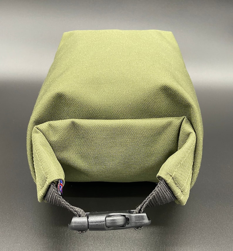 May include: Olive green rectangular pouch with a black buckle closure. The pouch is made of a textured fabric and features a small flag detail on the strap.