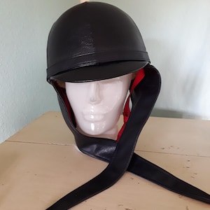 May include: A black equestrian-style helmet featuring a smooth, rounded outer shell with a short front brim. The interior lining is a vibrant red, and the helmet includes long, black, pointed side flaps that extend downward, secured by red straps.