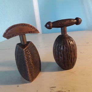 Op de afbeelding: Twee vintage notenkrakers, een met een bruine houten handgreep en een getextureerd bruin lichaam, de andere met een koperen lichaam en een bruine leren bovenkant.