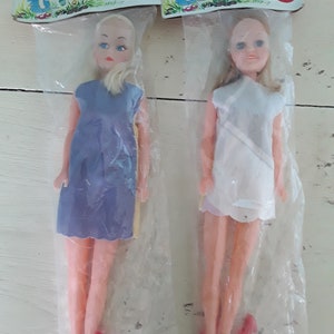 Op de afbeelding: Twee vintage Joelle poppen in plastic verpakking. De ene pop draagt een blauwe jurk, de andere een witte jurk. Beide poppen hebben blond haar, blauwe ogen en roze schoenen. De verpakking heeft een cartoon illustratie en de naam "Joelle".