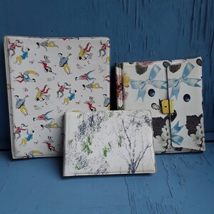 Puede incluir: Tres álbumes de fotos vintage con diferentes diseños. Un álbum tiene un fondo blanco con ilustraciones negras y rojas de personas bailando. Otro álbum tiene un fondo blanco con un diseño de árbol negro y verde. El tercer álbum tiene un fondo blanco con un diseño floral y una cinta azul.