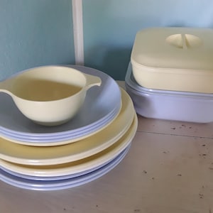 Può includere: Un set di stoviglie color pastello composto da una pila di piatti azzurri e giallo pallido, una piccola ciotola giallo pallido con manici laterali e un contenitore quadrato giallo pallido con coperchio sopra un contenitore quadrato azzurro.