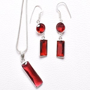 May include: A silver necklace and earrings set with red gemstone pendants. The necklace features a long, rectangular pendant and the earrings have a round gemstone at the top and a rectangular pendant below.