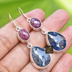 May include: A pair of silver dangle earrings with a teardrop-shaped blue gemstone and a smaller oval-shaped red gemstone. The earrings are on a hand.