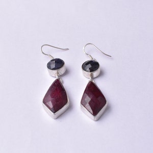 May include: A pair of silver dangle earrings with a dark blue gemstone and a red gemstone. The earrings have a teardrop shape.