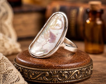 May include: A silver ring featuring an elongated, irregular-shaped white pearl with an iridescent luster. The pearl is set in a smooth silver bezel, and the ring band is made of polished silver, showcasing a clean and simple metal construction.