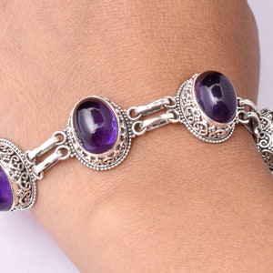 May include: A silver bracelet with three oval-shaped amethyst gemstones. The bracelet has a delicate chain design and intricate floral details.