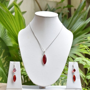 Op de afbeelding: Een zilveren ketting met een rode druppelvormige hanger en bijpassende oorbellen. De ketting en oorbellen worden gepresenteerd op een witte standaard.