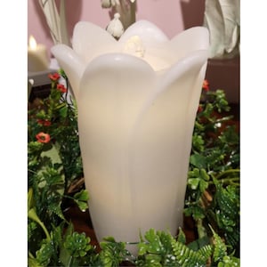 May include: A large, white, tulip-shaped candle with a soft, glowing interior. The candle is surrounded by green foliage and small red flowers, creating a natural, decorative display. The candle's petals are gently curved, giving it a realistic appearance.