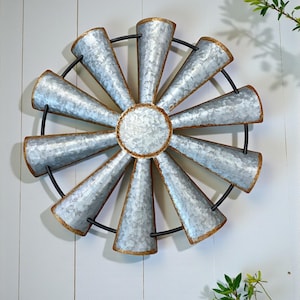May include: A galvanized metal windmill wall decor with a rustic design. The windmill features twelve blades with a silver finish and gold-colored edges, arranged around a central circle. A black metal ring connects the blades. The decor is mounted on a white wooden wall.