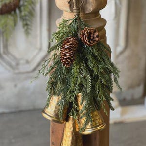 Puede incluir: Un adorno colgante decorativo que presenta tres campanas de metal martillado en tono dorado. Las campanas están adornadas con ramas de cedro artificiales verdes y dos piñas marrones, todo suspendido de un lazo marrón oscuro en la parte superior.