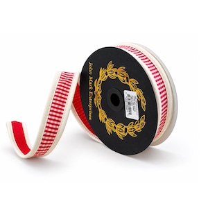 May include: A spool of ribbon featuring a red and white gingham pattern on one side and a solid red fabric on the other, bordered by cream-colored piping. The black spool label displays the text John Mark Enterprises and a gold laurel wreath.